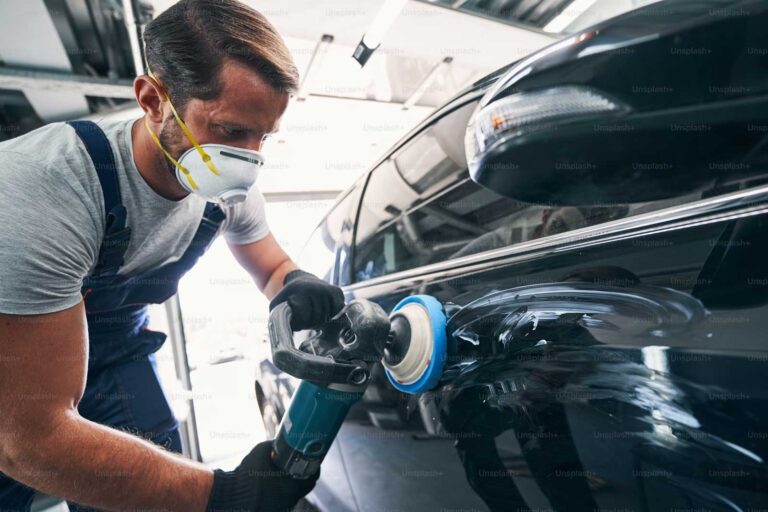 Technician wearing a mask and gloves buffing a black car's body panel with a rotary polisher