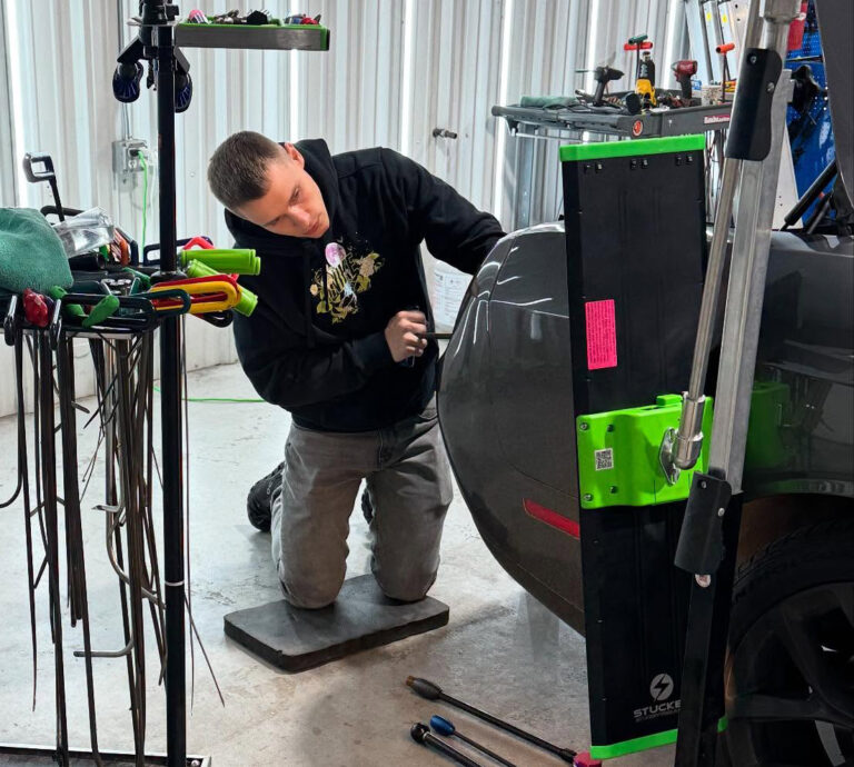 Technician kneeling beside a car, using a PDR tool to repair a dent under bright work light in a Houston shop