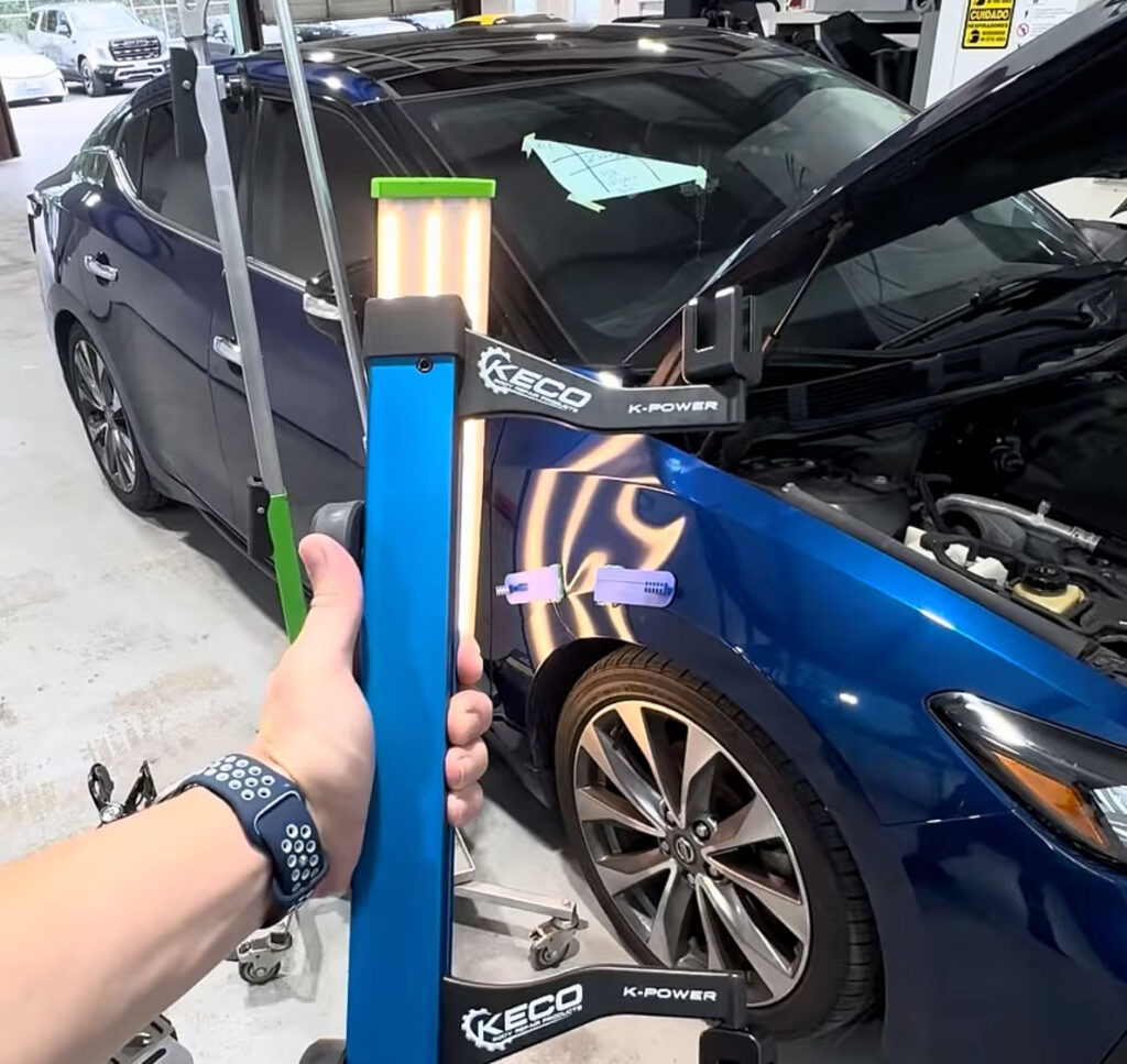 A technician uses a KECO lateral tension bar and cold glue pulling techniques on a silver Nissan Maxima fender to repair a large dent without using paint or Bondo.
