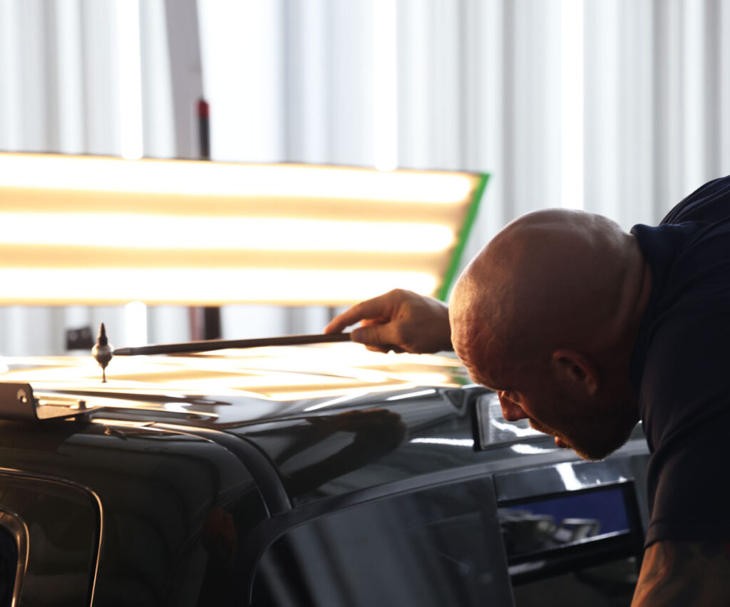 American Dent Specialists technician using a paintless dent repair tool to inspect a car's roof under fluorescent lighting