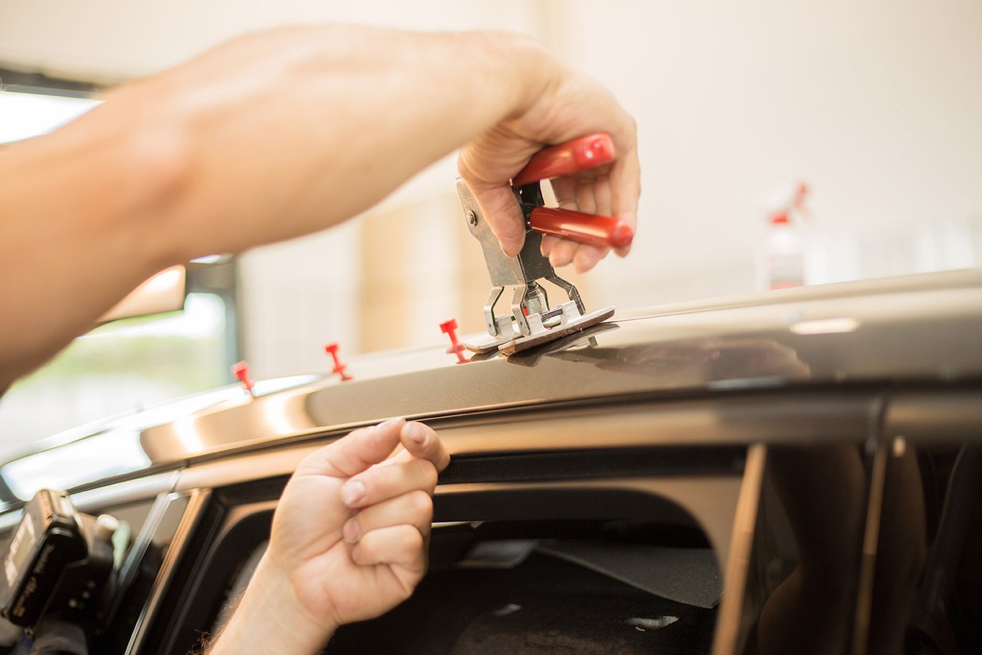Technician using a glue-pull tool with red tabs to lift a dent from a car roof edge